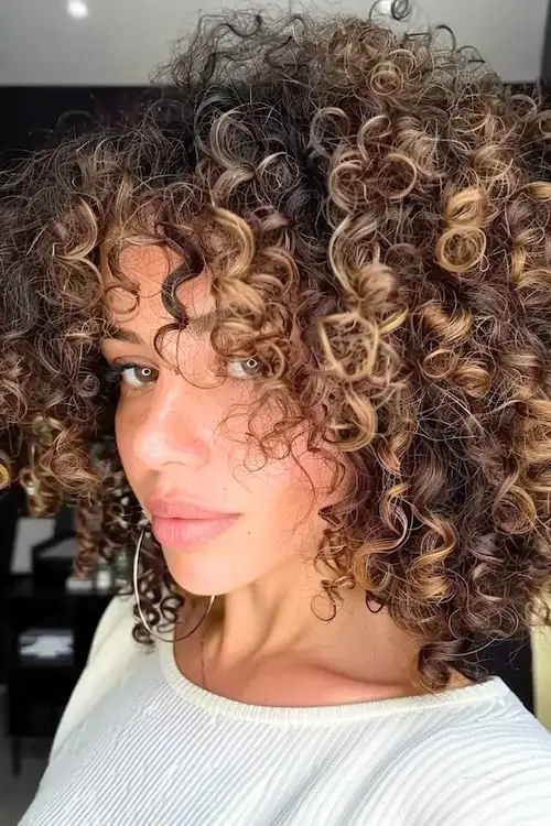 Tight curly dark brown hair with caramel blonde highlights enhancing the curls